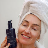 Woman in a towel turban smiling with eyes closed holding Mukti Hydrating Cleansing Lotion