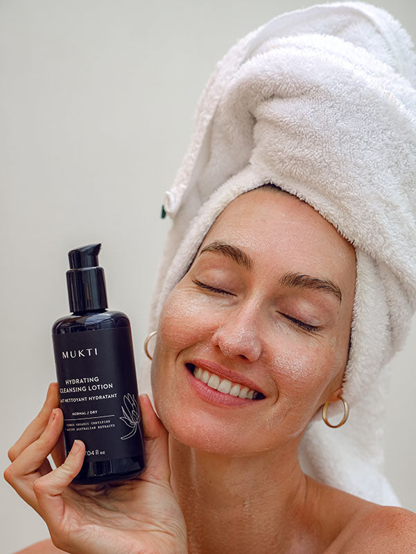 Woman in a towel turban smiling with eyes closed holding Mukti Hydrating Cleansing Lotion