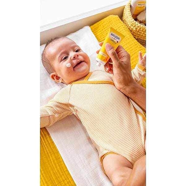Smiling baby wearing yellow, lying down and being applied Calendula Face Cream on his face.