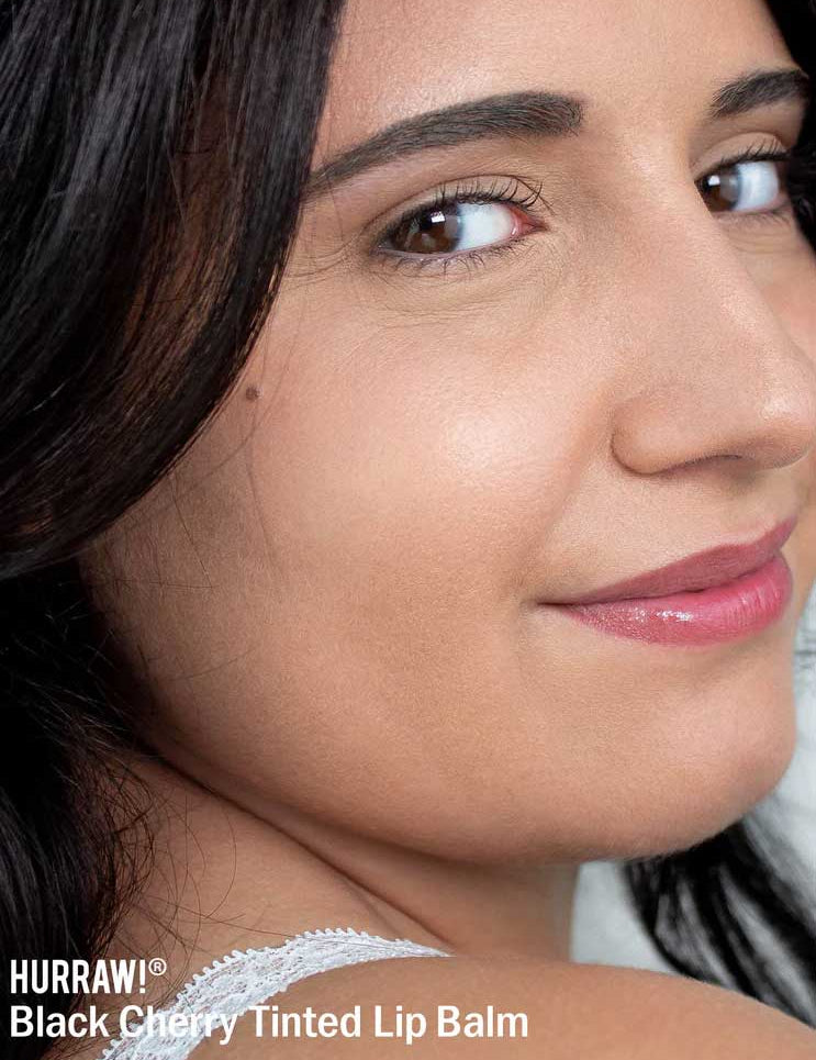 A close-up of a woman with smooth skin, dark hair, and glossy tinted lips, smiling slightly. The text below reads: Hurraw! Organic Lip Balm - Black Cherry 4.8g for naturally beautiful lips.