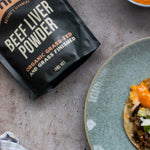 Chief Beef Liver Powder pouch beside a blue-grey plate holding a round tortilla topped with browned filling, chopped vegetables and orange sauce.