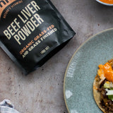 Chief Beef Liver Powder pouch beside a blue-grey plate holding a round tortilla topped with browned filling, chopped vegetables and orange sauce.