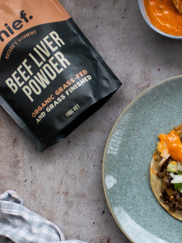 Chief Beef Liver Powder pouch beside a blue-grey plate holding a round tortilla topped with browned filling, chopped vegetables and orange sauce.