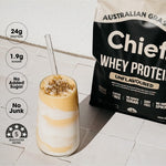 Tall layered pale yellow and white shake topped with granola beside a Chief Unflavoured Whey Protein pouch. Graphic badges state 24g protein, 1.9g sugar, no added sugar, no junk and a five-star health rating.