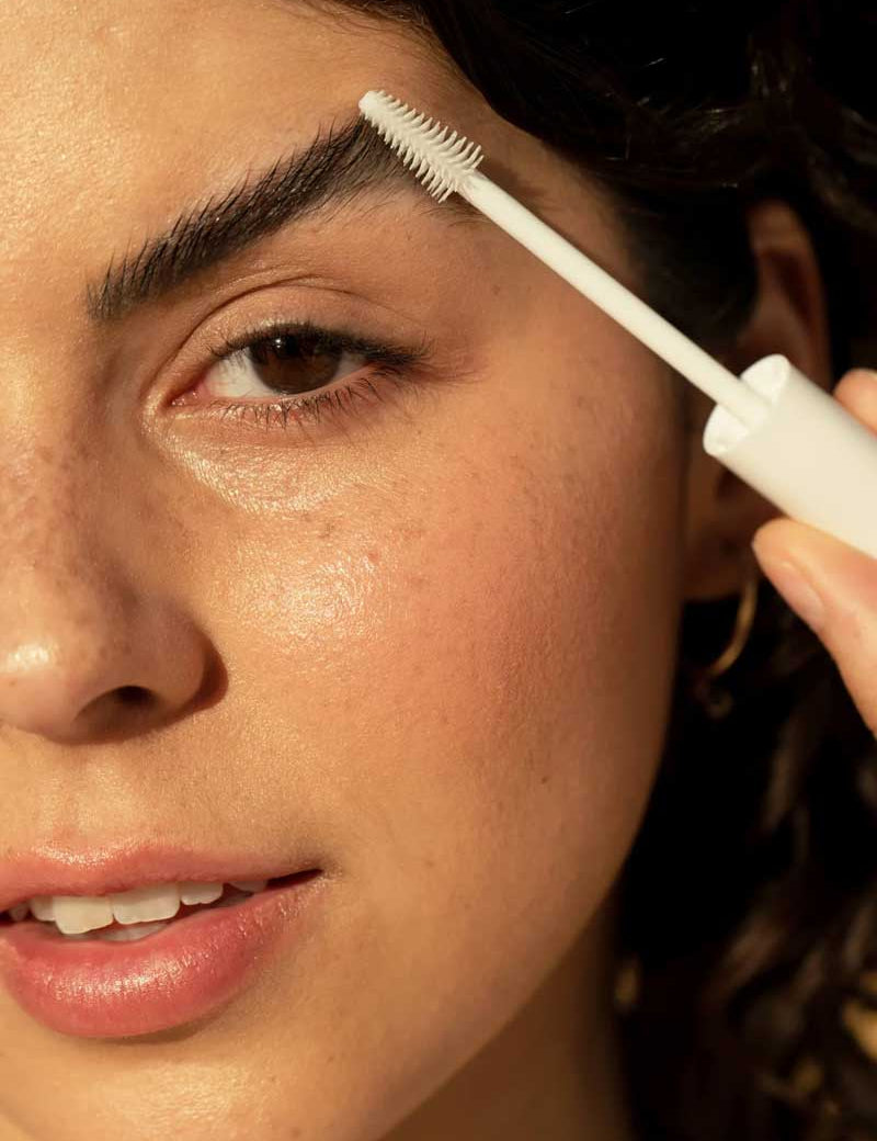 A woman grooms her dark eyebrows with the Ere Perez Aloe Gel Lash & Brow Mascara - Clear 10ml, using a white brush. Her skin is clear and glowing, lips slightly parted, and freckles visible.