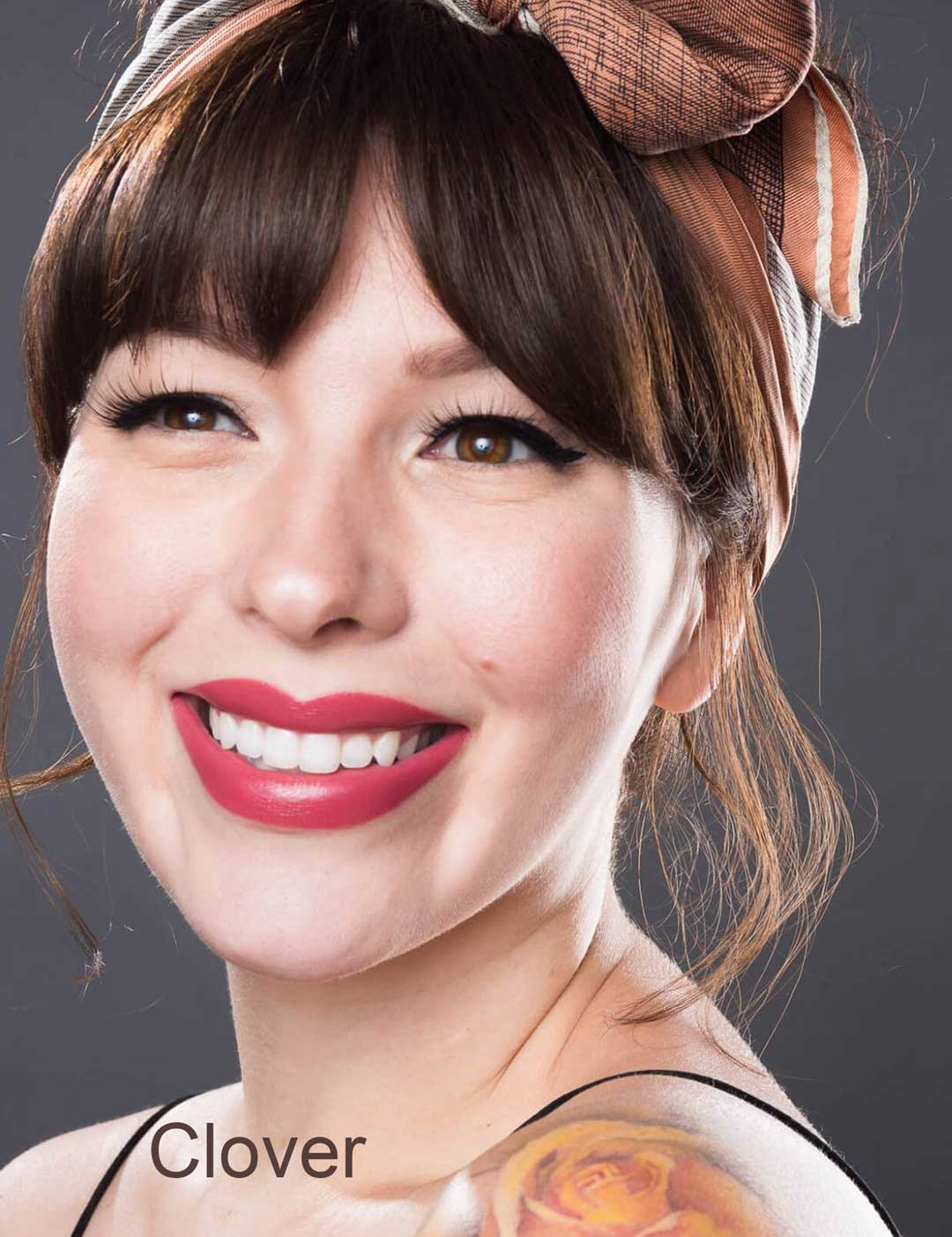 A young woman with brown hair and bangs smiles brightly, wearing winged eyeliner, a patterned headscarf, a colorful shoulder tattoo, and 100% Pure Fruit Pigmented® Pomegranate Oil Moisturising Lipstick. "Clover" appears in the bottom left corner.
