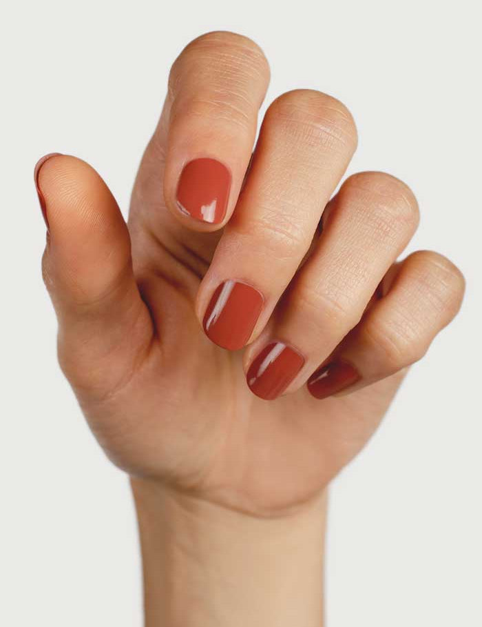 A hand with fingers slightly curled displays neatly manicured nails painted in Sienna Byron Bay Nail Polish - Courage, a glossy terracotta shade, against a plain light background.