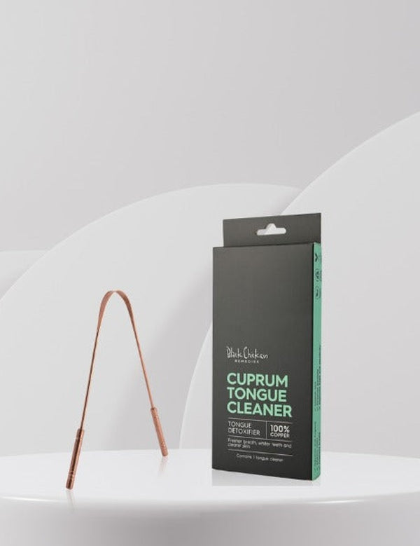 U-shaped copper tongue scraper standing beside its tall black and mint-green carton on a white ledge against a pale grey background.