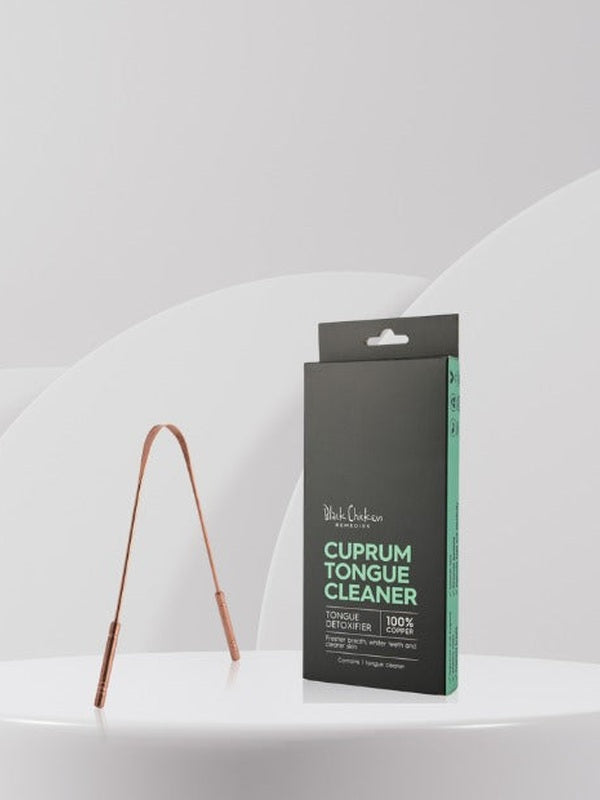 U-shaped copper tongue scraper standing beside its tall black and mint-green carton on a white ledge against a pale grey background.