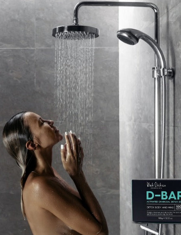 A woman stands beneath a running rainfall shower with her face tilted upwards and palms near her chin. A black D-Bar carton is pictured at the lower right.