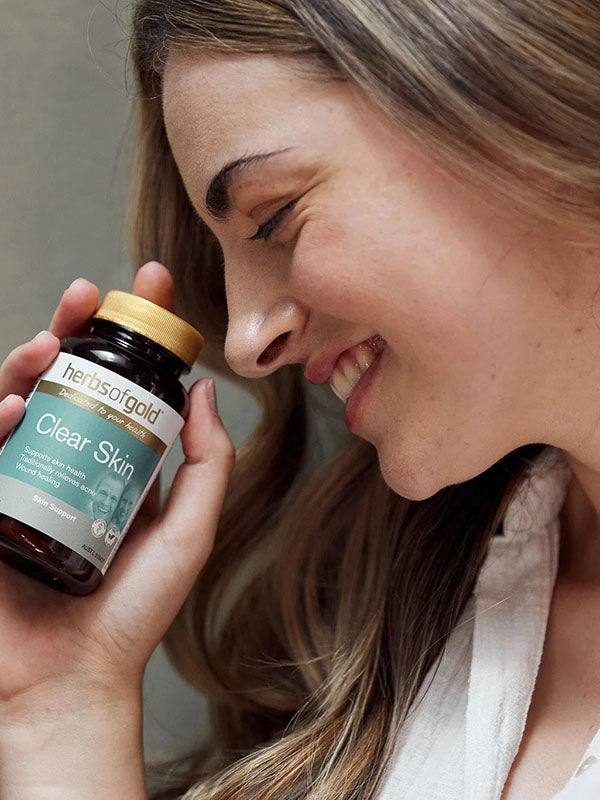 Smiling woman holding the Herbs of Gold Clear Skin bottle beside her cheek
