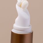 Extreme close-up of the curved white applicator on an INIKA Eye Cream tube, with a small bead of pale cream emerging from its opening above a gold collar.