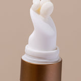 Extreme close-up of the curved white applicator on an INIKA Eye Cream tube, with a small bead of pale cream emerging from its opening above a gold collar.