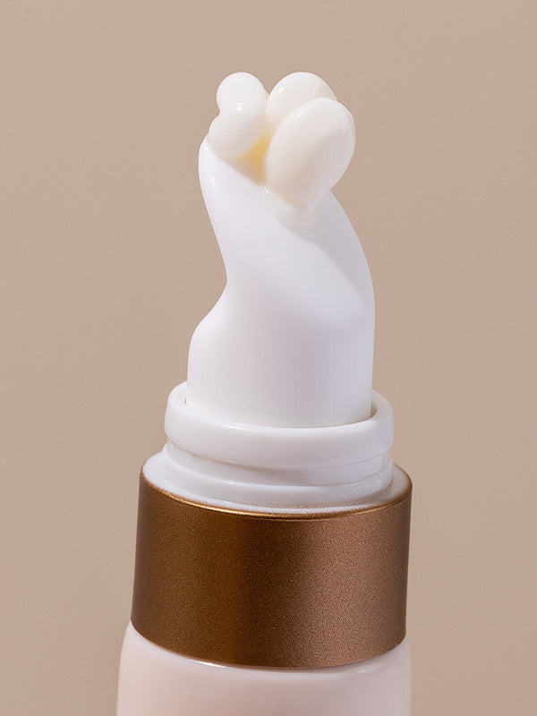 Extreme close-up of the curved white applicator on an INIKA Eye Cream tube, with a small bead of pale cream emerging from its opening above a gold collar.