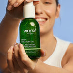 A smiling young woman holding a green bottle of Weleda Deep Cleansing Gel in front of her, with a blue background.