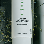 Close-up of a tall white Madara Deep Moisture Night Cream bottle with a green branching motif and black label text, surrounded by fine green stems and watery reflections.