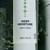 Close-up of a tall white Madara Deep Moisture Night Cream bottle with a green branching motif and black label text, surrounded by fine green stems and watery reflections.