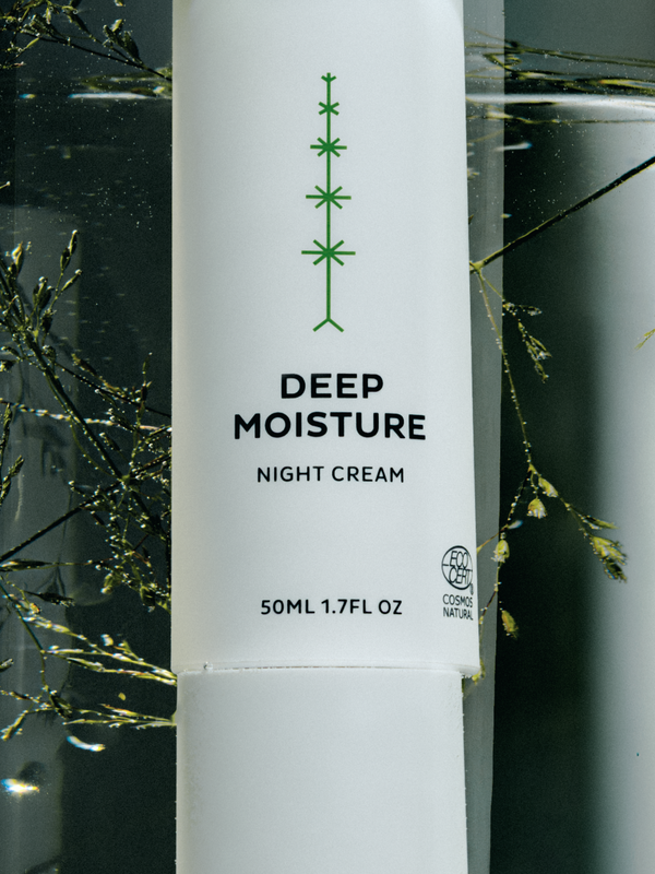 Close-up of a tall white Madara Deep Moisture Night Cream bottle with a green branching motif and black label text, surrounded by fine green stems and watery reflections.