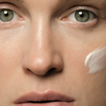 Close-up of a woman's face with pale green eyes and a thick white dab of cream on one cheek.