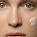 Close-up of a woman's face with pale green eyes and a thick white dab of cream on one cheek.