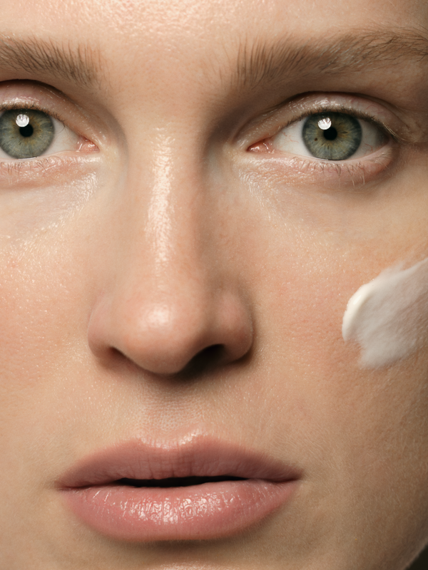 Close-up of a woman's face with pale green eyes and a thick white dab of cream on one cheek.