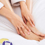 Person in a white robe rubbing balm onto their ankle while sitting on white bedding, with an open tin of yellow Dr. Bronner's Arnica-Menthol Magic Balm and its lid beside their feet.