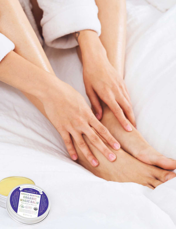 Person in a white robe rubbing balm onto their ankle while sitting on white bedding, with an open tin of yellow Dr. Bronner's Arnica-Menthol Magic Balm and its lid beside their feet.