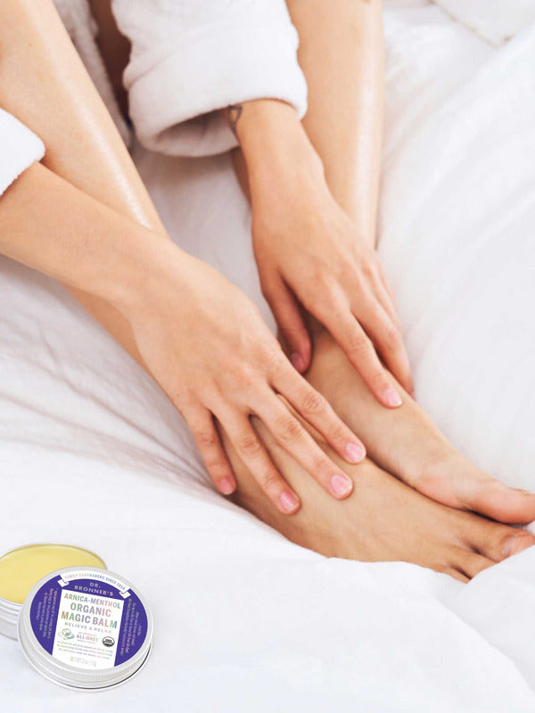 Person in a white robe rubbing balm onto their ankle while sitting on white bedding, with an open tin of yellow Dr. Bronner's Arnica-Menthol Magic Balm and its lid beside their feet.