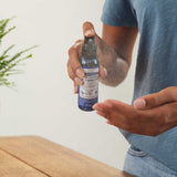 Person in a blue T-shirt spraying Dr. Bronner's Peppermint Organic Hand Sanitiser toward an open palm above a wooden counter.