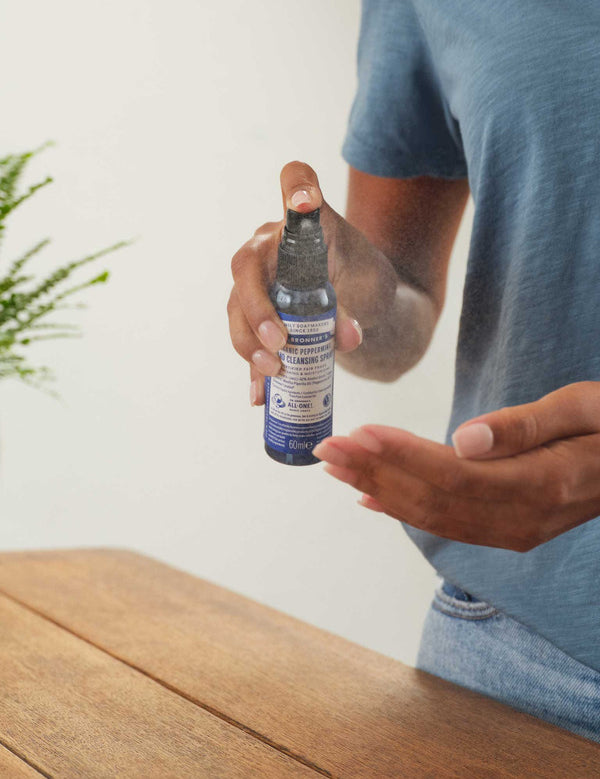 Person in a blue T-shirt spraying Dr. Bronner's Peppermint Organic Hand Sanitiser toward an open palm above a wooden counter.