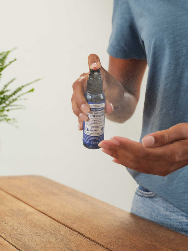Person in a blue T-shirt spraying Dr. Bronner's Peppermint Organic Hand Sanitiser toward an open palm above a wooden counter.