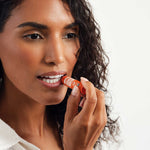 Curly-haired woman in a white top applying Dr. Bronner's Orange Ginger Lip Balm to her lower lip from an orange tube.