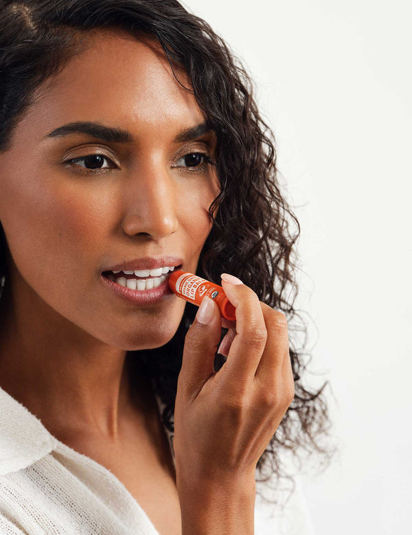 Curly-haired woman in a white top applying Dr. Bronner's Orange Ginger Lip Balm to her lower lip from an orange tube.