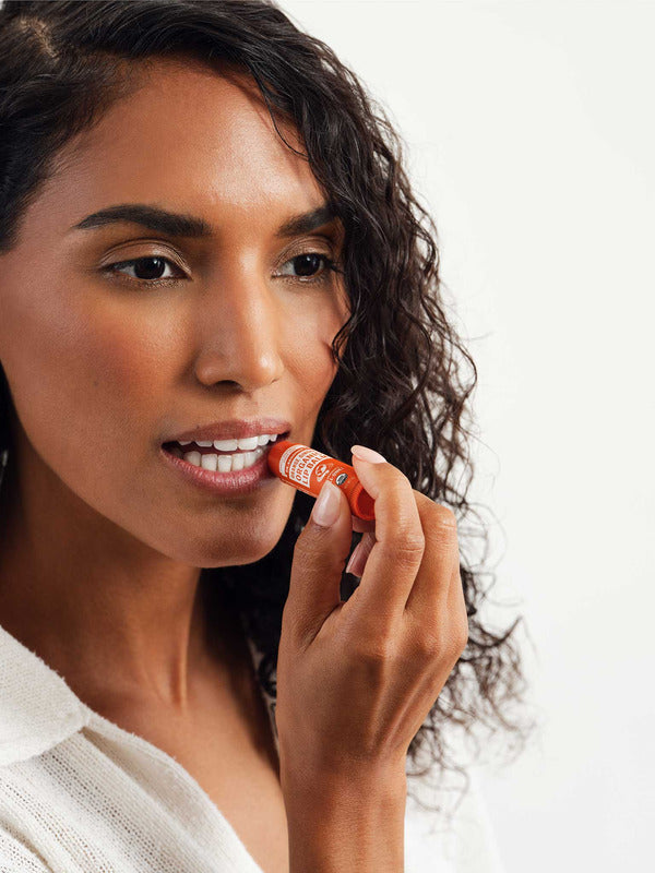 Curly-haired woman in a white top applying Dr. Bronner's Orange Ginger Lip Balm to her lower lip from an orange tube.