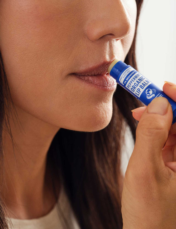 Close-up of a woman applying Dr. Bronner's Peppermint Lip Balm from a dark blue tube to the edge of her lips.