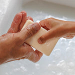 Close-up of wet hands holding a rectangular ivory soap bar above a white basin filled with bubbly water.