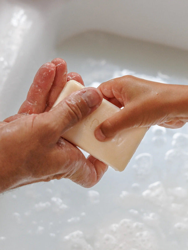 Close-up of wet hands holding a rectangular ivory soap bar above a white basin filled with bubbly water.
