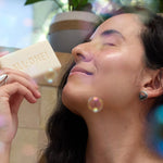 Woman with closed eyes holding an ivory Dr. Bronner's soap bar embossed ALL-ONE! near her nose, with floating bubbles and leafy plants around her.