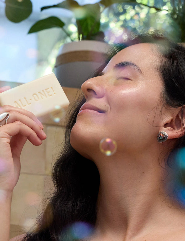 Woman with closed eyes holding an ivory Dr. Bronner's soap bar embossed ALL-ONE! near her nose, with floating bubbles and leafy plants around her.