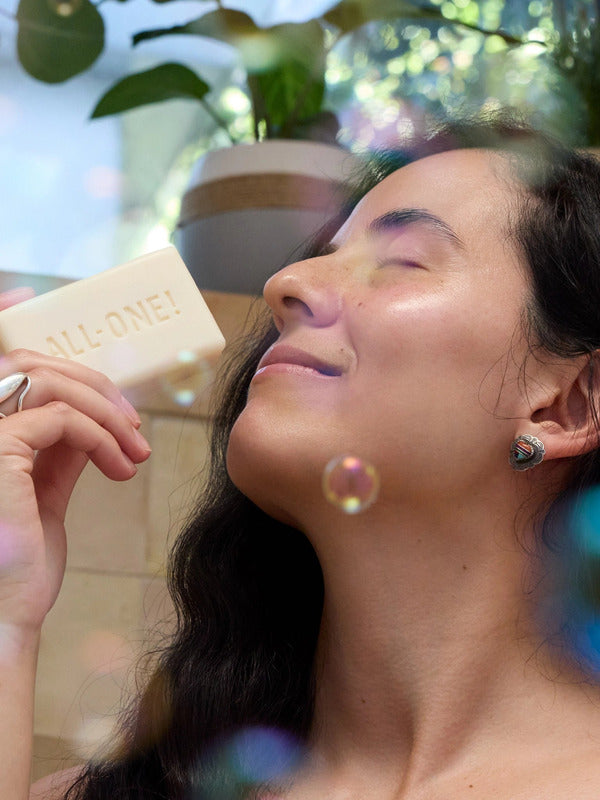 Woman with closed eyes holding an ivory Dr. Bronner's soap bar embossed ALL-ONE! near her nose, with floating bubbles and leafy plants around her.