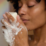 Person with short curly hair and closed eyes holding an ivory soap bar near their nose, with thick white lather covering their hands.