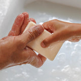 Close-up of wet hands holding a rectangular ivory soap bar above a white basin filled with bubbly water.