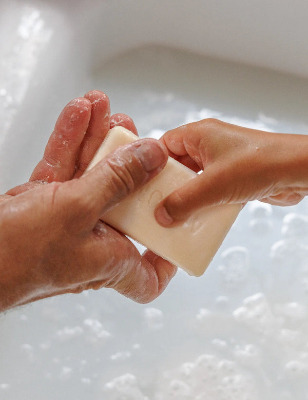 Close-up of wet hands holding a rectangular ivory soap bar above a white basin filled with bubbly water.