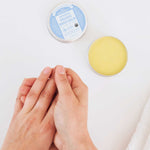 Overhead view of hands rubbing together beside an open tin of yellow Dr. Bronner's Unscented Magic Balm and its blue-labelled lid on a white surface.