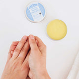 Overhead view of hands rubbing together beside an open tin of yellow Dr. Bronner's Unscented Magic Balm and its blue-labelled lid on a white surface.