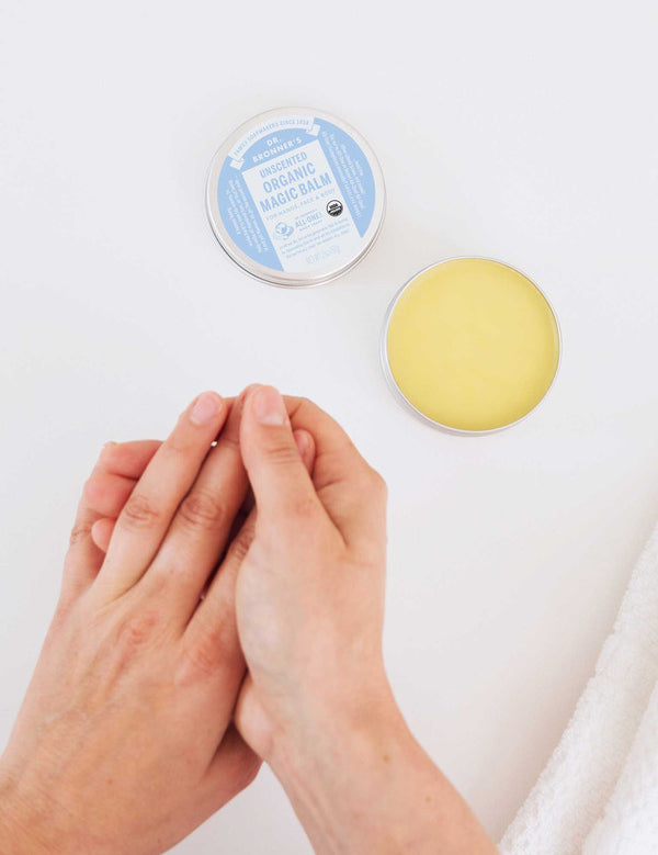 Overhead view of hands rubbing together beside an open tin of yellow Dr. Bronner's Unscented Magic Balm and its blue-labelled lid on a white surface.
