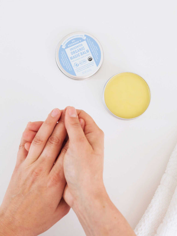Overhead view of hands rubbing together beside an open tin of yellow Dr. Bronner's Unscented Magic Balm and its blue-labelled lid on a white surface.