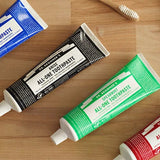 Dr. Bronner's All-One Toothpaste tubes arranged on a wooden counter, with the black Anise tube featured among Peppermint, Spearmint and Cinnamon.