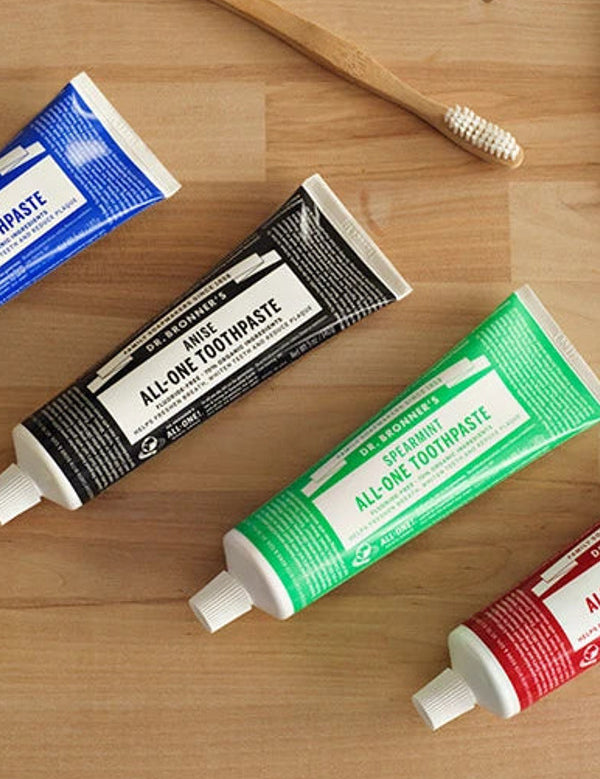 Dr. Bronner's All-One Toothpaste tubes arranged on a wooden counter, with the black Anise tube featured among Peppermint, Spearmint and Cinnamon.