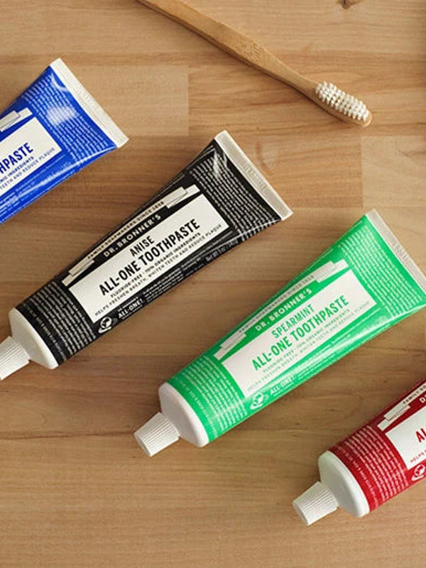 Dr. Bronner's All-One Toothpaste tubes arranged on a wooden counter, with the black Anise tube featured among Peppermint, Spearmint and Cinnamon.
